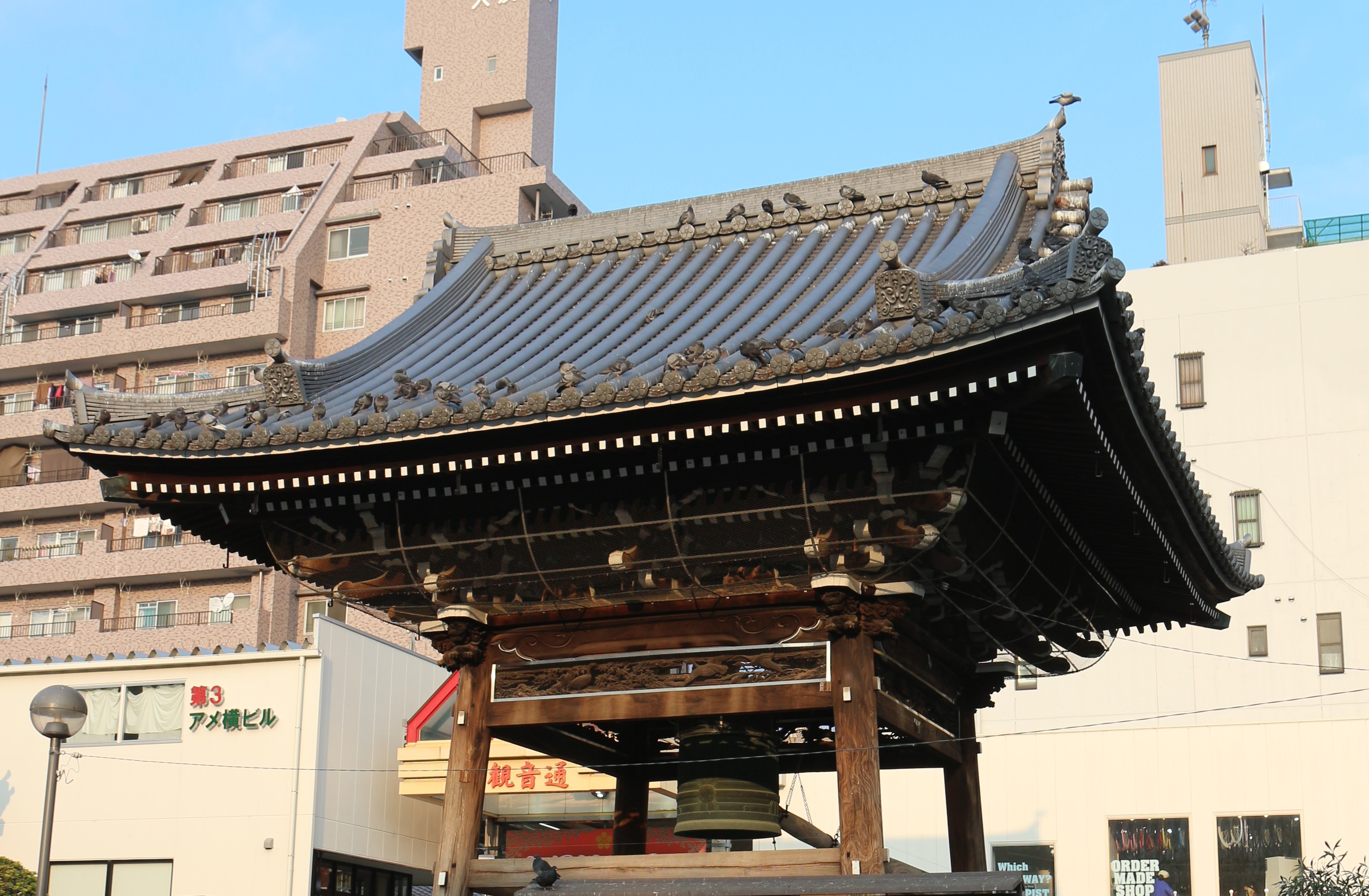 Photo de la petite annexe à côté de Osu Kanon. On aperçoit la cloche rituelle, et de nombreux pigeons sont posés au bord du toit incurvé.