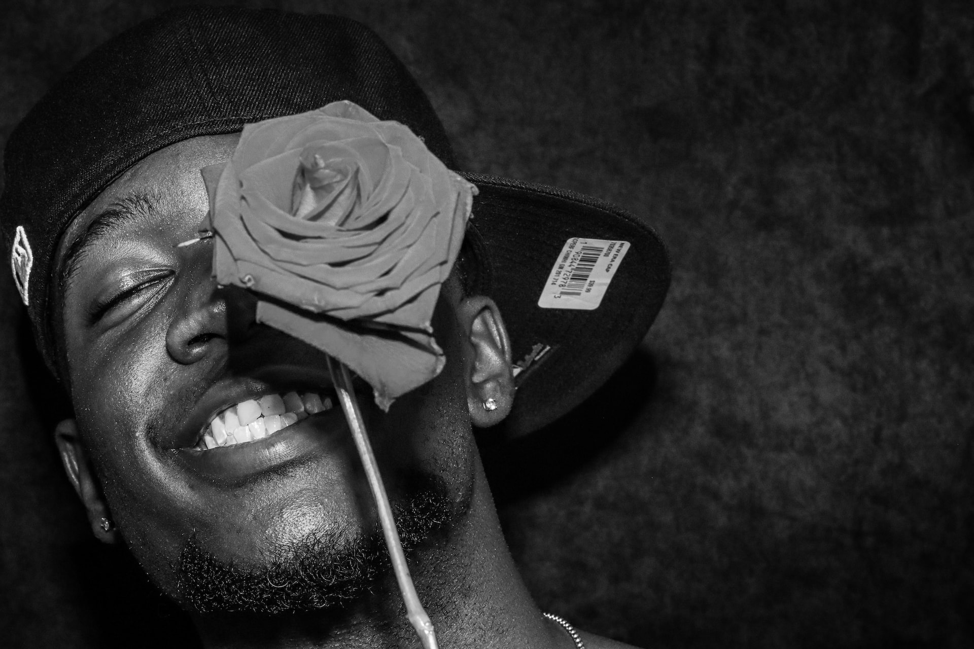 Homme souriant portant une casquette dont on voit encore l'étiquette. Son visage est à moitié caché derrière une rose bon marché.