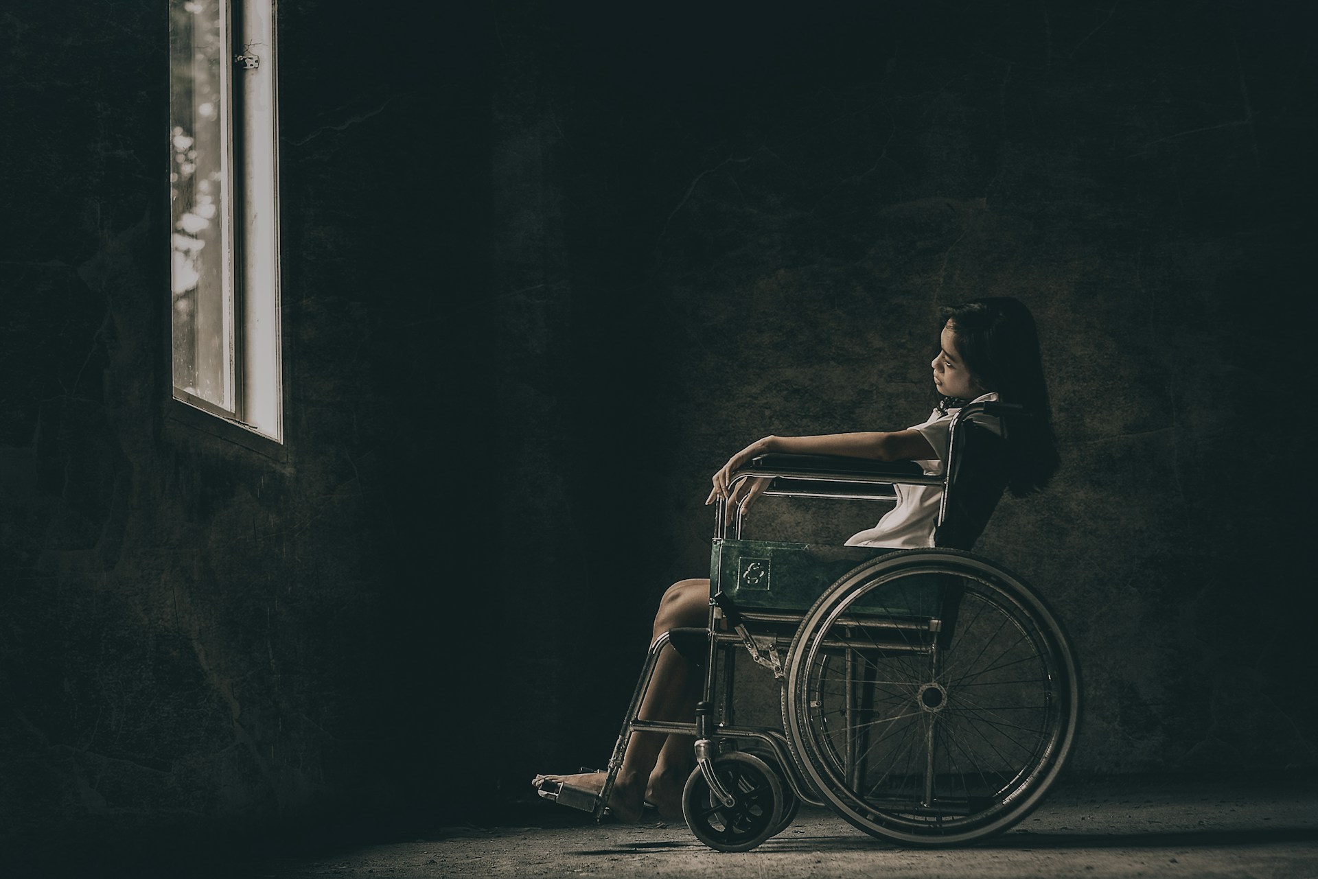 Jeune fille assise dans un fauteuil roulant, la tête penchée, nostalgique et dépitée, face à une fenêtre ouverte sur l'extérieur vers lequel elle ne peut plus courir.