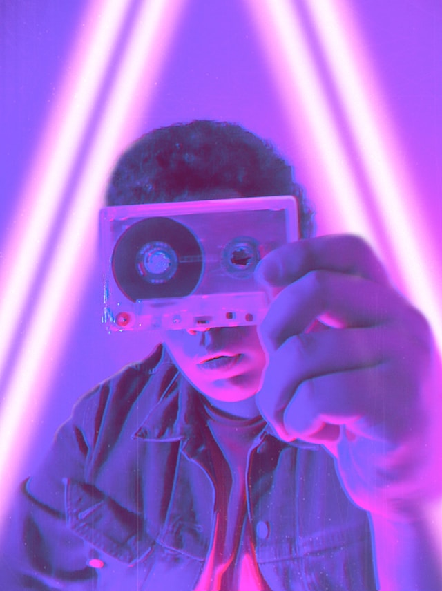 Dans un halo de couleur violette fluorescente, un jeune homme tient, devant son visage, une cassette audio des années 80