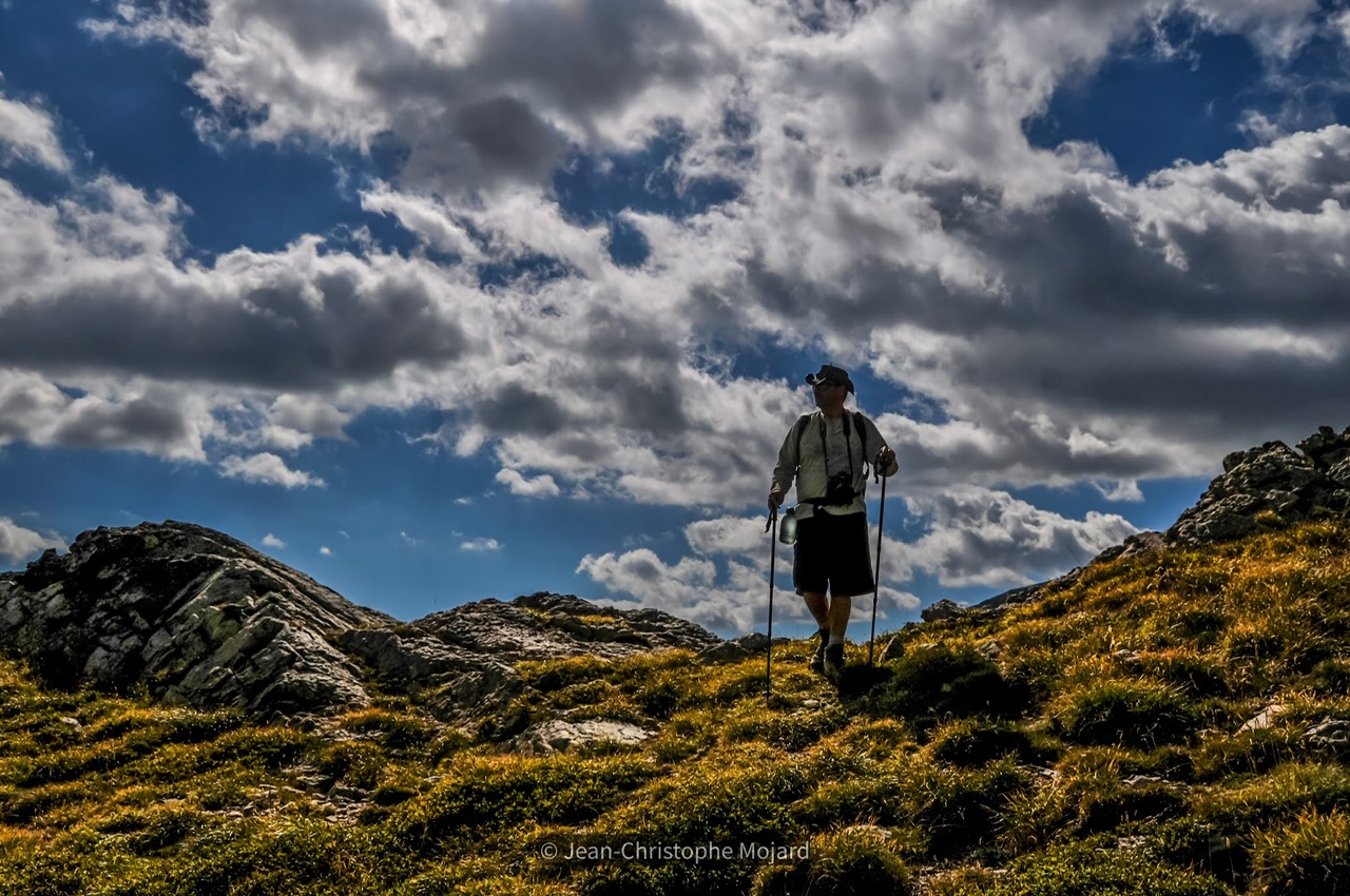 Photo personnelle où je franchis un col montagneux en Corse, à la végétation rase sous un ciel bleu profond parsemé de nuages blancs.